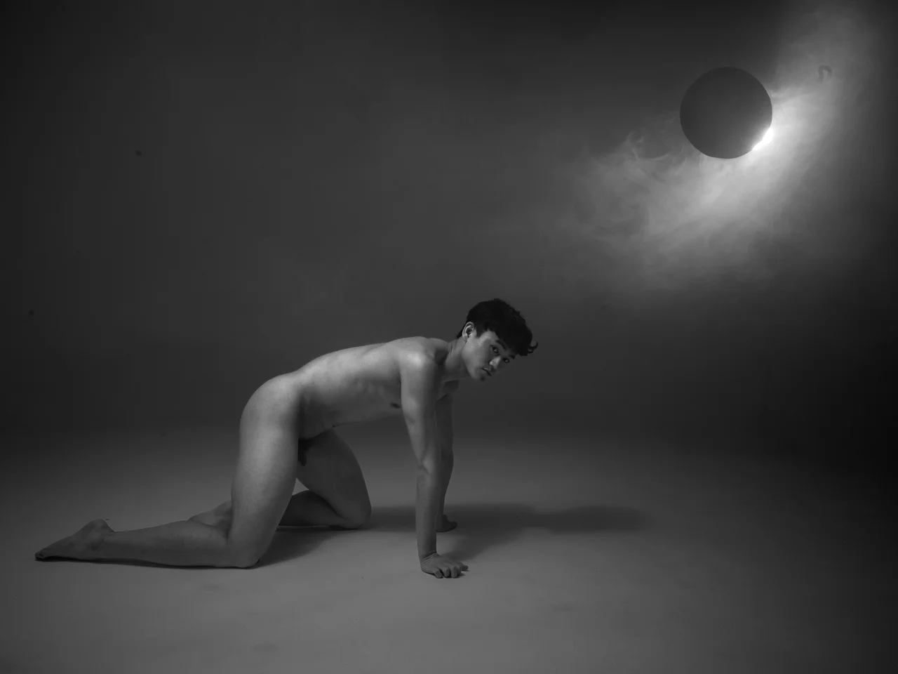 Black-and-white portrait of a kneeling person under dramatic light with an art ball above, minimalist composition